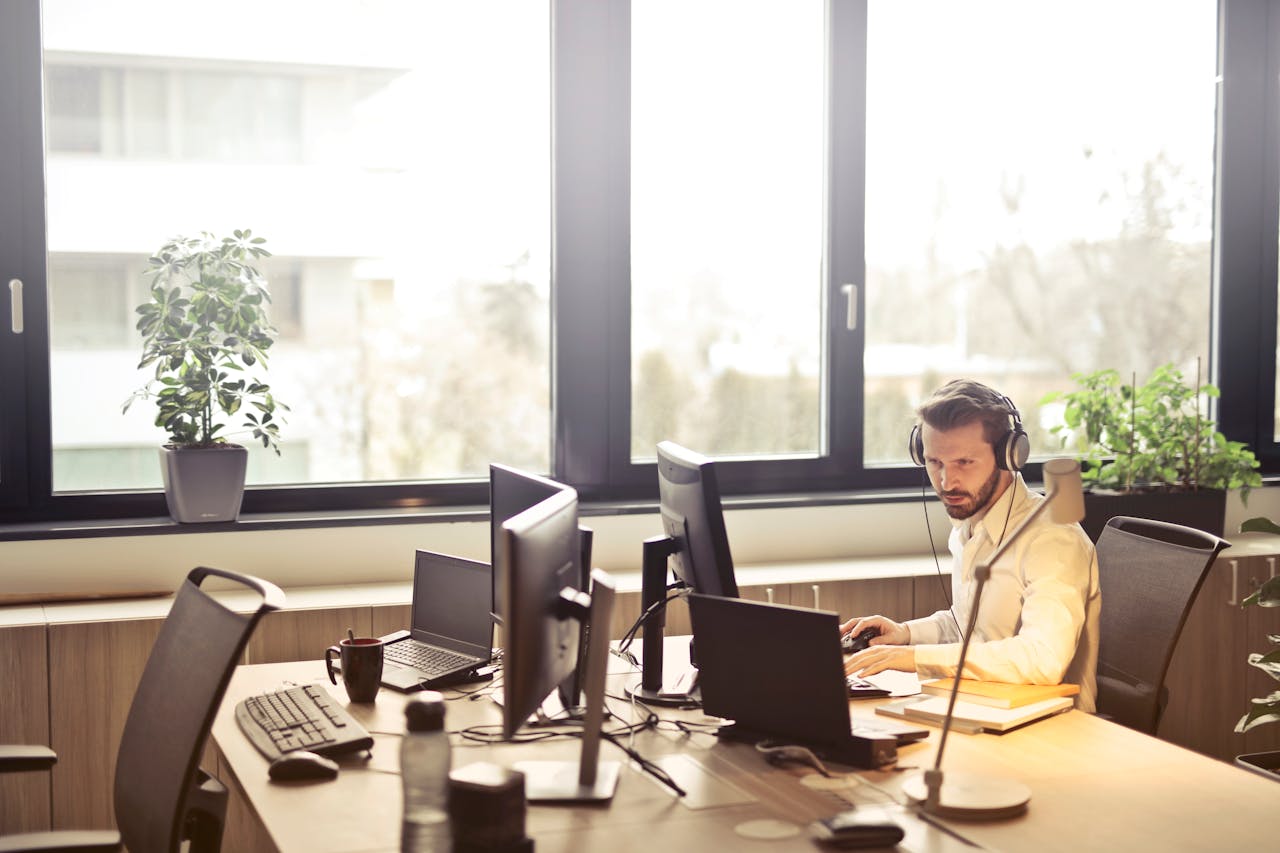 services-02 A businessman sits at a desk using multiple computers and a headset in a well-lit modern office.